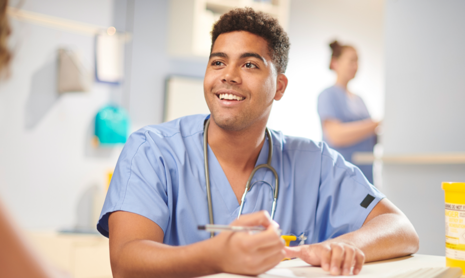 A man is wearing hospital scrubs with a stethoscope around his neck. He smiles to someone off camera while writing on paper.